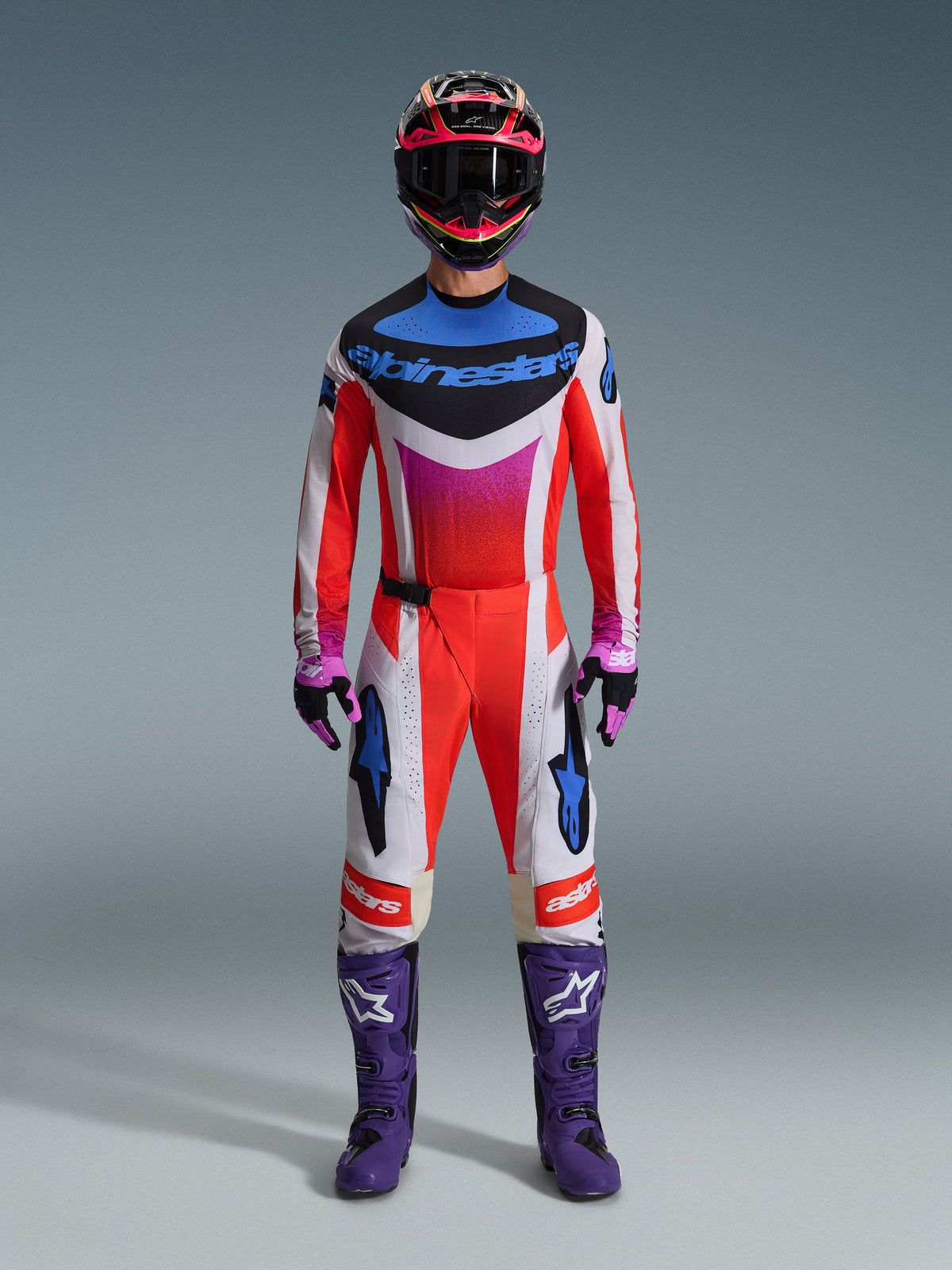 Pilote de motocross portant un équipement de course complet, maillot Alpinestars Techstar Knif 2026 et pantalon assorti en blanc et orange avec des accents bleus et violets, bottes de course violettes avec logo blanc et casque multicolore, tenue coordonnée pour la compétition de course tout-terrain