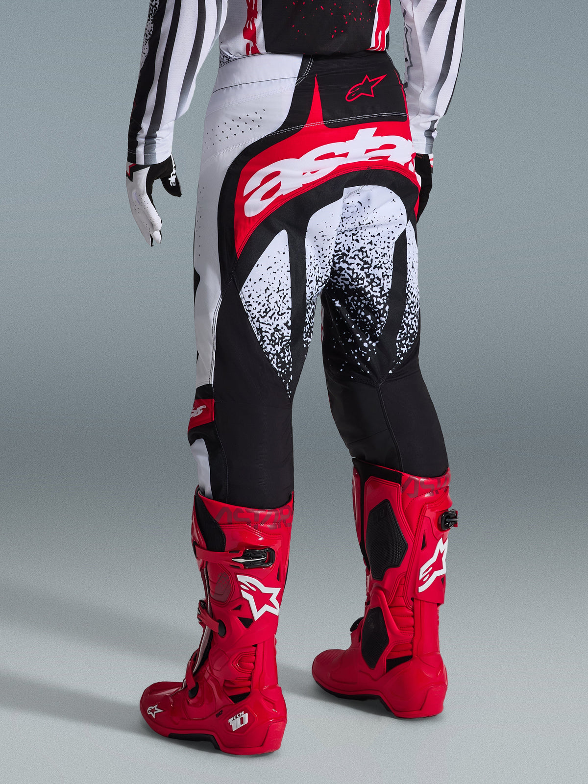 Alpinestars 2026 Techstar Nomur Hose, Motocross-Hose, Schwarz, Weiß und Rot, Rückansicht an einem Fahrer mit passenden roten Stiefeln, mit markantem rot-weißem „astars“-Logo auf der Sitzfläche und gesprenkelten Grafikmustern, professionelle Offroad-Rennausrüstung