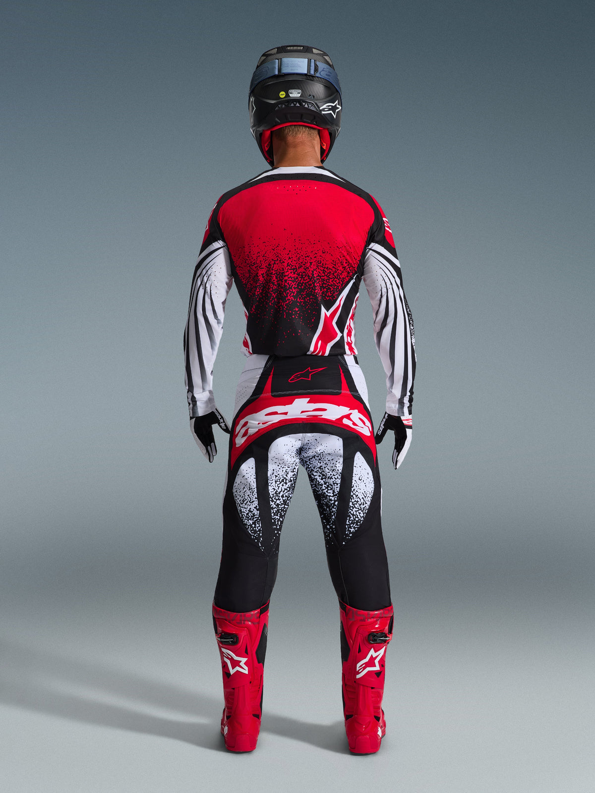 Rückansicht eines Motocross-Fahrers in der Alpinestars 2026 Techstar Nomur Hose in Schwarz-Weiß-Rot, mit markantem Branding am Gesäß und gesprenkelten Grafiken, kombiniert mit einem passenden roten Trikot und leuchtend roten Stiefeln für eine komplette Offroad-Rennausrüstung.