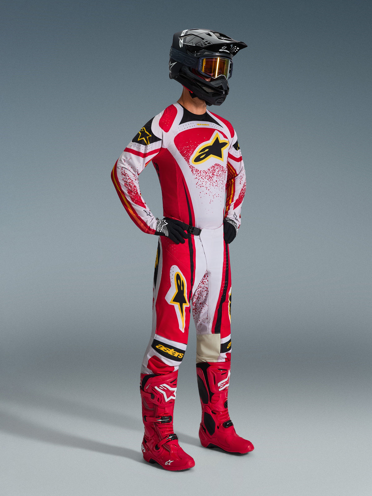 Pilote de motocross portant l'ensemble d'équipement Alpinestars Techstar Nomur 2026, rouge et blanc avec accents noirs et logos jaunes, porté avec des bottes rouges et un casque noir assortis, tenue de course professionnelle pour la compétition tout-terrain