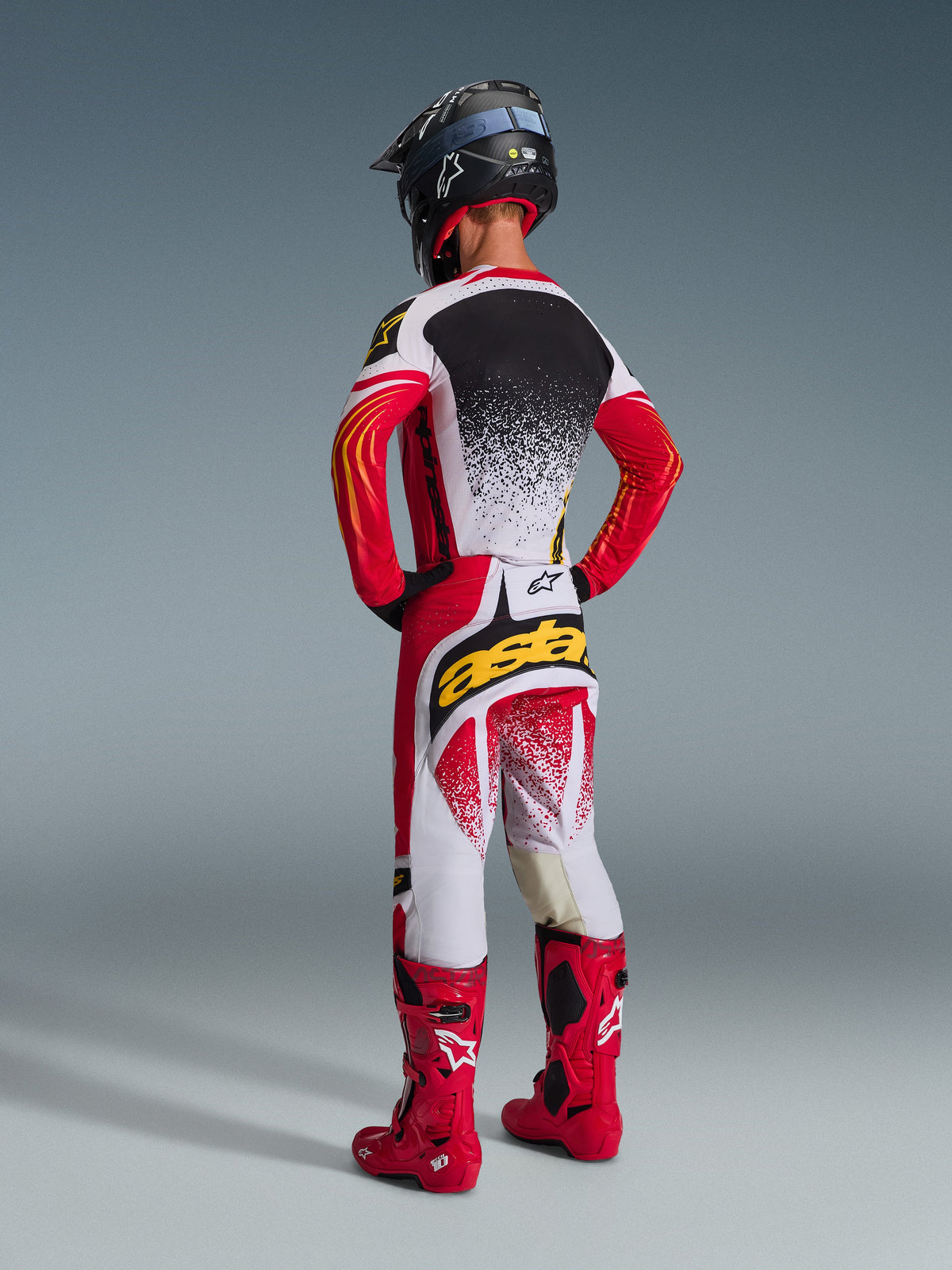 Pilote de motocross de dos portant la tenue Alpinestars 2026 Techstar Nomur, maillot rouge, blanc et noir avec dos moucheté blanc, pantalon assorti avec logo 'astars' jaune sur l'assise, bottes de course rouge vif et casque noir, tenue de motocross complète et coordonnée