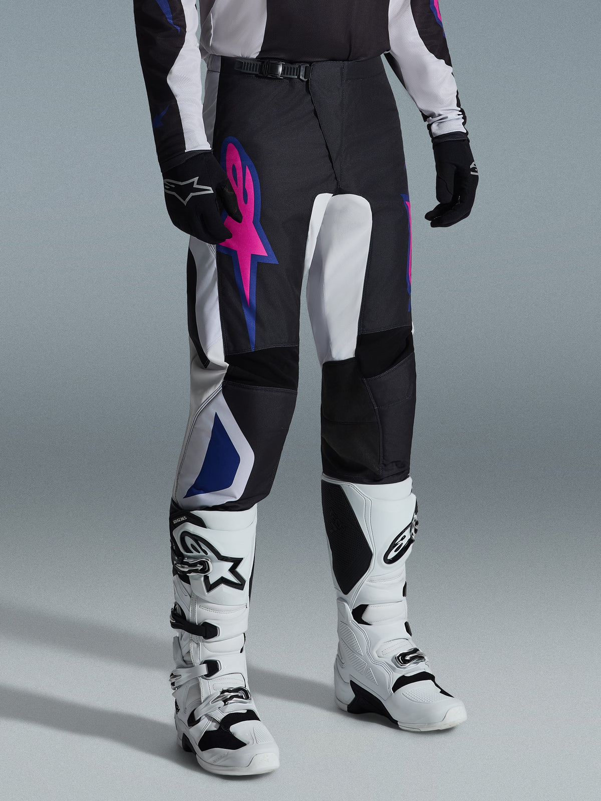 Alpinestars 2026 Fluid Grid Pants, pantalon de motocross, noir, gris clair et violet, avec un logo étoile rose vif et des accents géométriques bleus, porté avec des bottes de motocross blanches et des gants noirs, équipement de conduite tout-terrain ergonomique