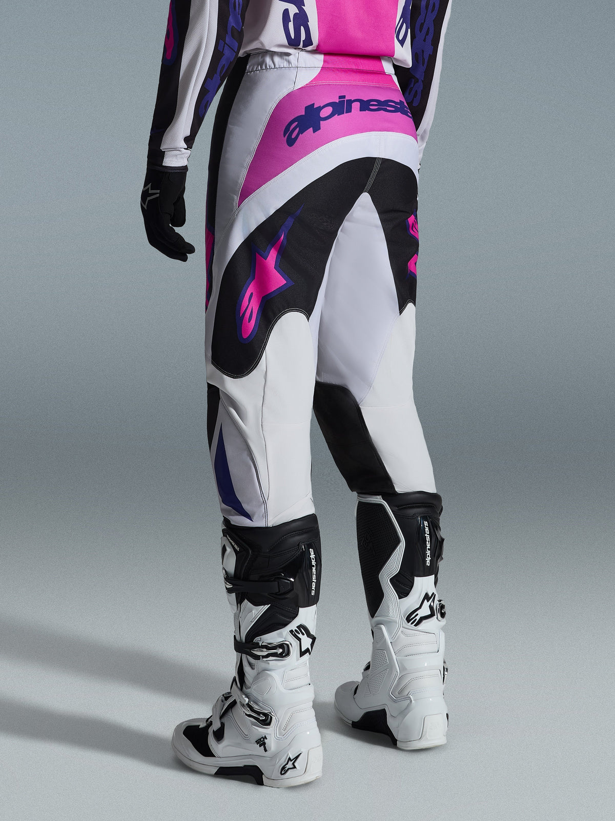 Pantalon Alpinestars Fluid Grid 2026, pantalon de motocross, noir, gris clair et violet, vue arrière du pilote portant le pantalon avec des bottes blanches et noires, avec des logos Alpinestars roses et violets, coupe athlétique pour la course tout-terrain