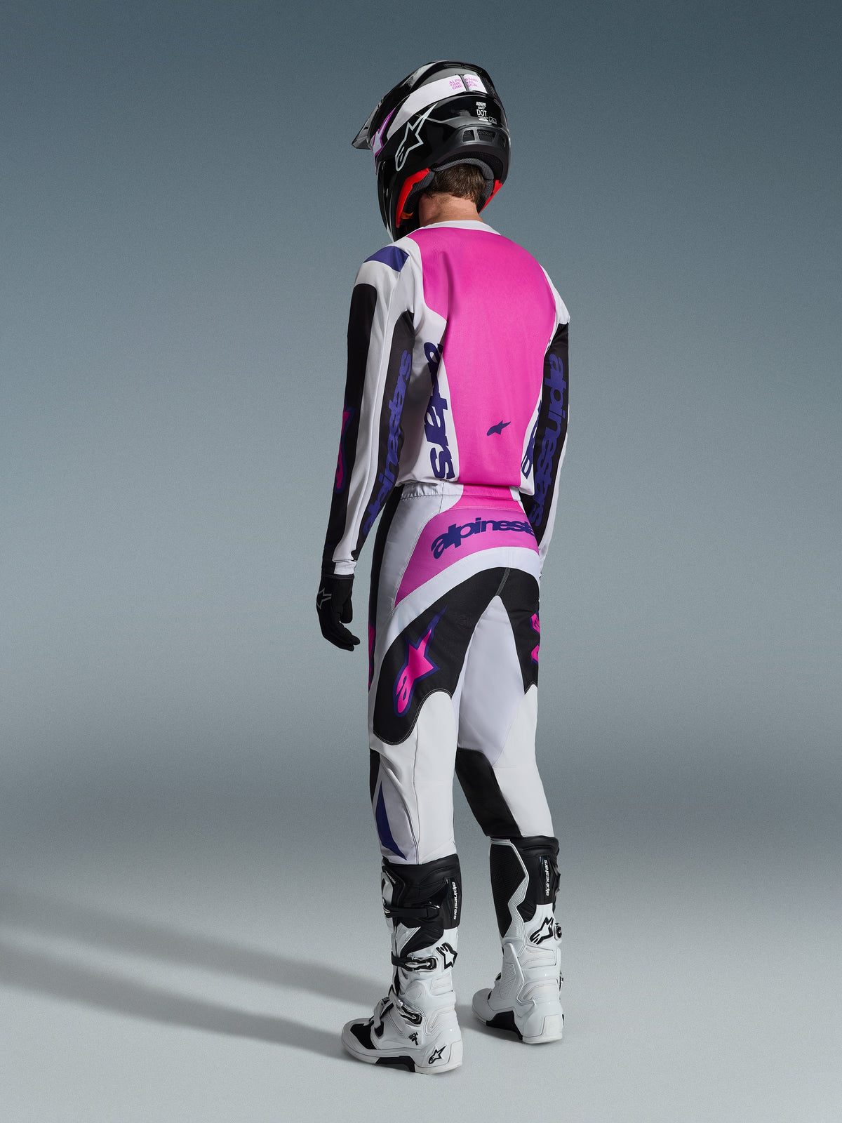 Alpinestars 2026 Fluid Grid Pants, pantalon de motocross, noir, gris clair et violet, vue arrière d'un pilote avec maillot assorti et bottes blanches, logos violets et panneaux rose vif, conçu pour la performance en motocross tout-terrain