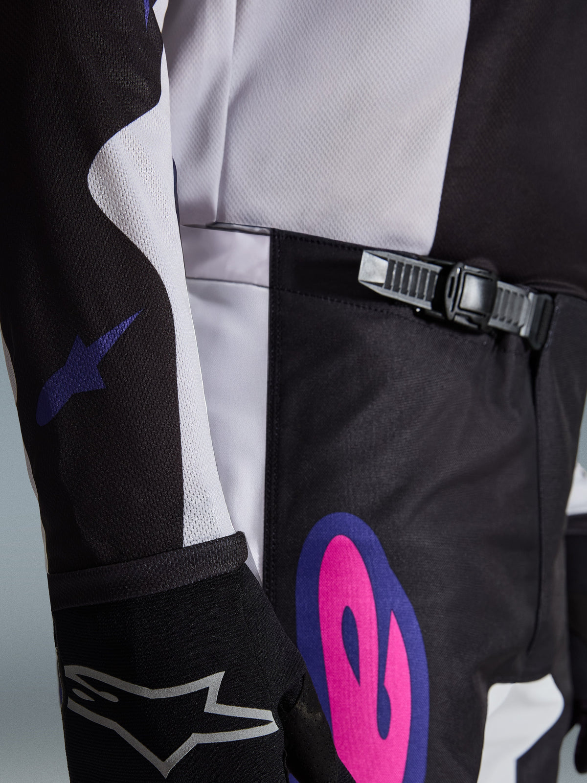 Alpinestars 2026 Fluid Grid Pants, pantalon de motocross, noir, gris clair et violet, gros-plan présentant le système de fermeture à cliquet à la taille et un graphisme rose et violet vif sur la hanche, porté avec un maillot assorti et un gant noir.