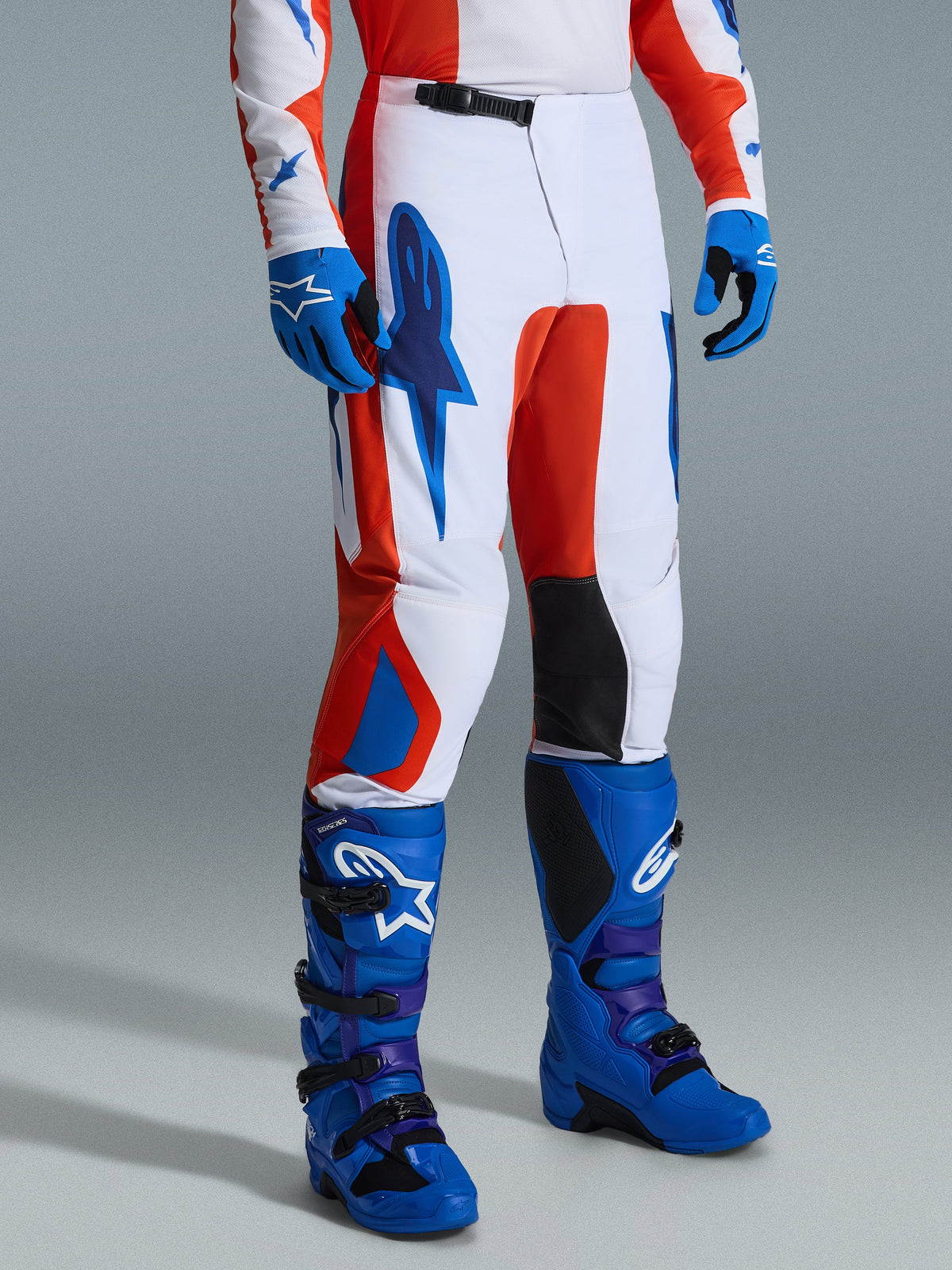 Alpinestars 2026 Fluid Grid Pants, pantalon de motocross, orange et bleu UCLA, porté avec des gants et des bottes bleus assortis, présentant une base blanche avec un marquage graphique audacieux, des panneaux intérieurs aux genoux noirs et une conception ergonomique pour le pilotage tout-terrain