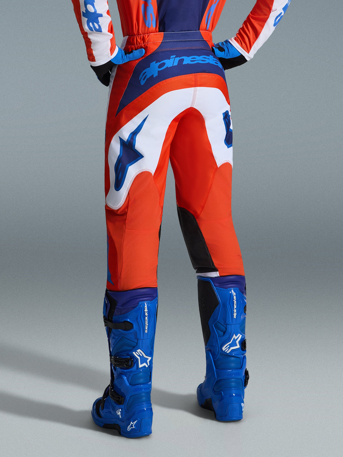 Alpinestars 2026 Fluid Grid Pants, pantalon de motocross, orange et bleu UCLA avec empiècements blancs, vue arrière montrant le marquage bleu vif sur l'assise et le logo étoilé, associé à des bottes de motocross bleues assorties, équipement de course tout-terrain professionnel