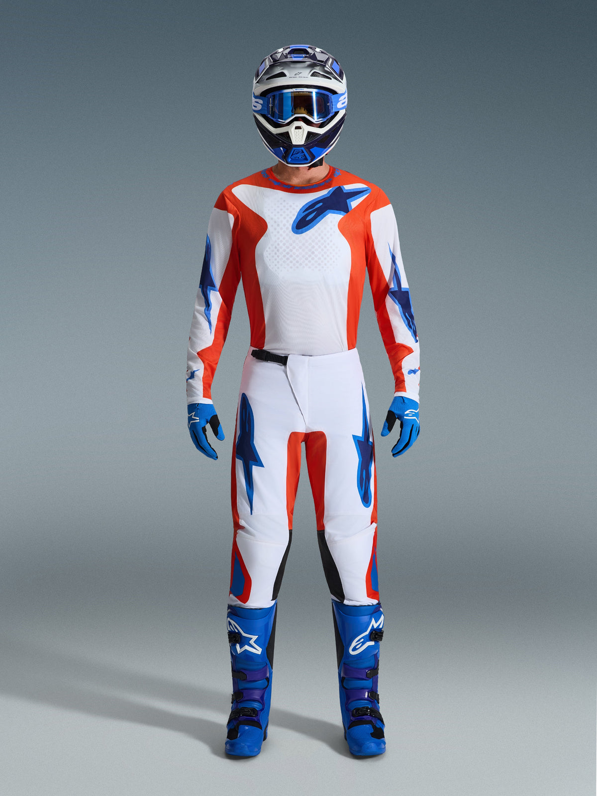 Pilote de motocross portant un équipement de course complet, Pantalon Alpinestars Fluid Grid 2026 en Orange et UCLA Blue, maillot blanc avec manches orange et logos bleus, casque et lunettes bleus assortis, bottes et gants bleus, tenue coordonnée pour la course tout-terrain