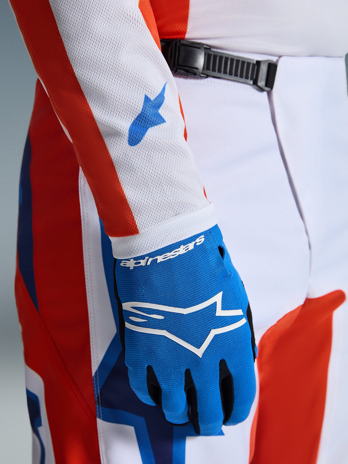 Pantalon Alpinestars Fluid Grid 2026, pantalon de motocross, orange et bleu UCLA, gros plan présentant un gant bleu assorti et une manche de maillot en mesh blanc avec des logos bleus, illustrant la fermeture à cliquet à la taille et la coupe athlétique.