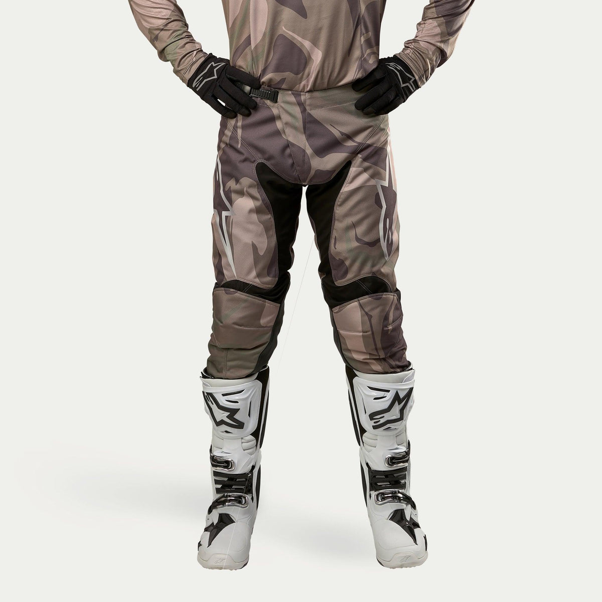 Alpinestars 2024 Racer Tactical broek, motorcrossbroek, Military Green Camo Brown, met grote witte Alpinestars-logo's op de dijbenen en zwarte panelen aan de binnenkant van de benen, gedragen met wit-zwarte motorcrosslaarzen en zwarte handschoenen, bijpassende camouflage jersey, vooraanzicht.