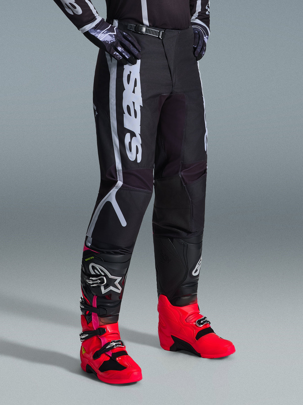 Pantalon Alpinestars Fluid Apex 2026, pantalon de motocross, noir et gris, porté avec des bottes rose vif et rouges et un maillot noir et blanc assorti, doté d'un marquage Alpinestars vertical sur la cuisse, de panneaux renforcés et d'une fermeture à cliquet à la taille pour une performance tout-terrain.