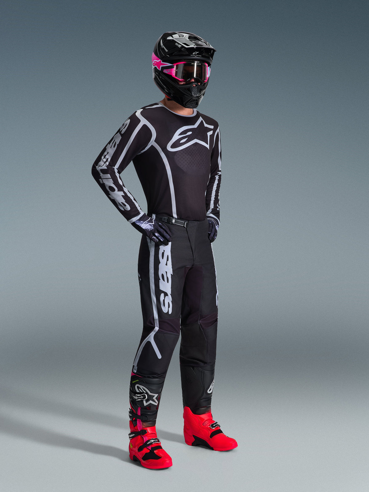 Alpinestars 2026 Fluid Apex Pants, pantalon de motocross, noir et gris, porté avec un maillot noir et blanc assorti, un casque noir avec des lunettes roses et des bottes rouge vif, avec un marquage blanc imposant sur les jambes et une coupe sport pour la course tout-terrain.