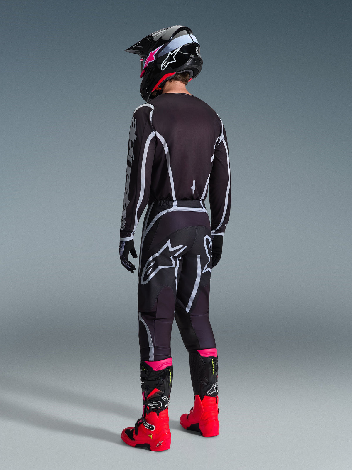 Pantalon Alpinestars Fluid Apex 2026, pantalon de motocross, noir et gris, vue arrière présentant le logo Alpinestars blanc sur l'assise, porté avec un maillot assorti, un casque noir et rose et des bottes rouge vif.