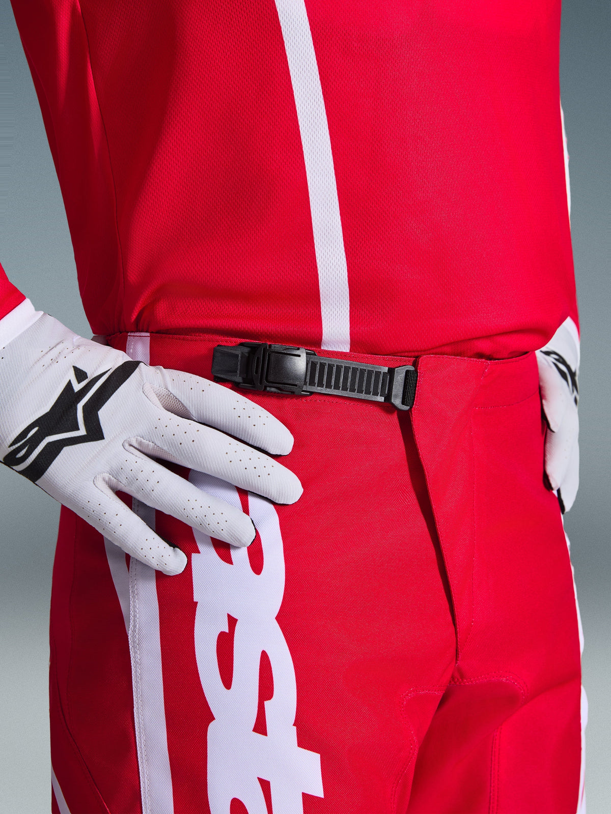Alpinestars 2026 Fluid Apex Hose, Motocross-Hose, Rot und Weiß, Nahaufnahme des schwarzen Ratschenverschlusses am Bund, getragen mit weißen Alpinestars Handschuhen und passendem rotem Jersey, strapazierfähige Performance-Ausrüstung für den Offroad-Sport.