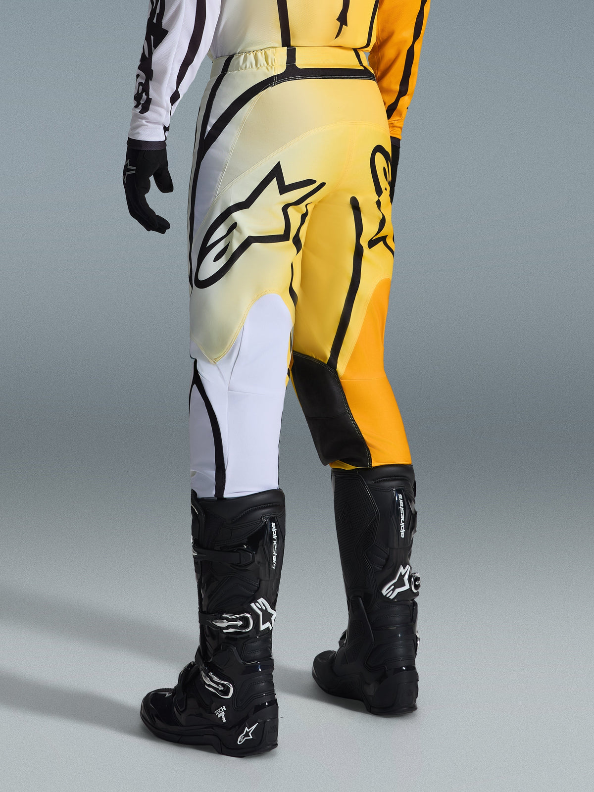 Alpinestars 2026 Fluid Apex broek, motorcross broek, geel en zwart kleurverloop met witte en oranje accenten, achteraanzicht, gedragen met bijpassende jersey en zwarte Tech 7 laarzen, voorzien van een opvallend zwart Alpinestars-logo op het zitvlak, technische uitrusting voor off-road rijden