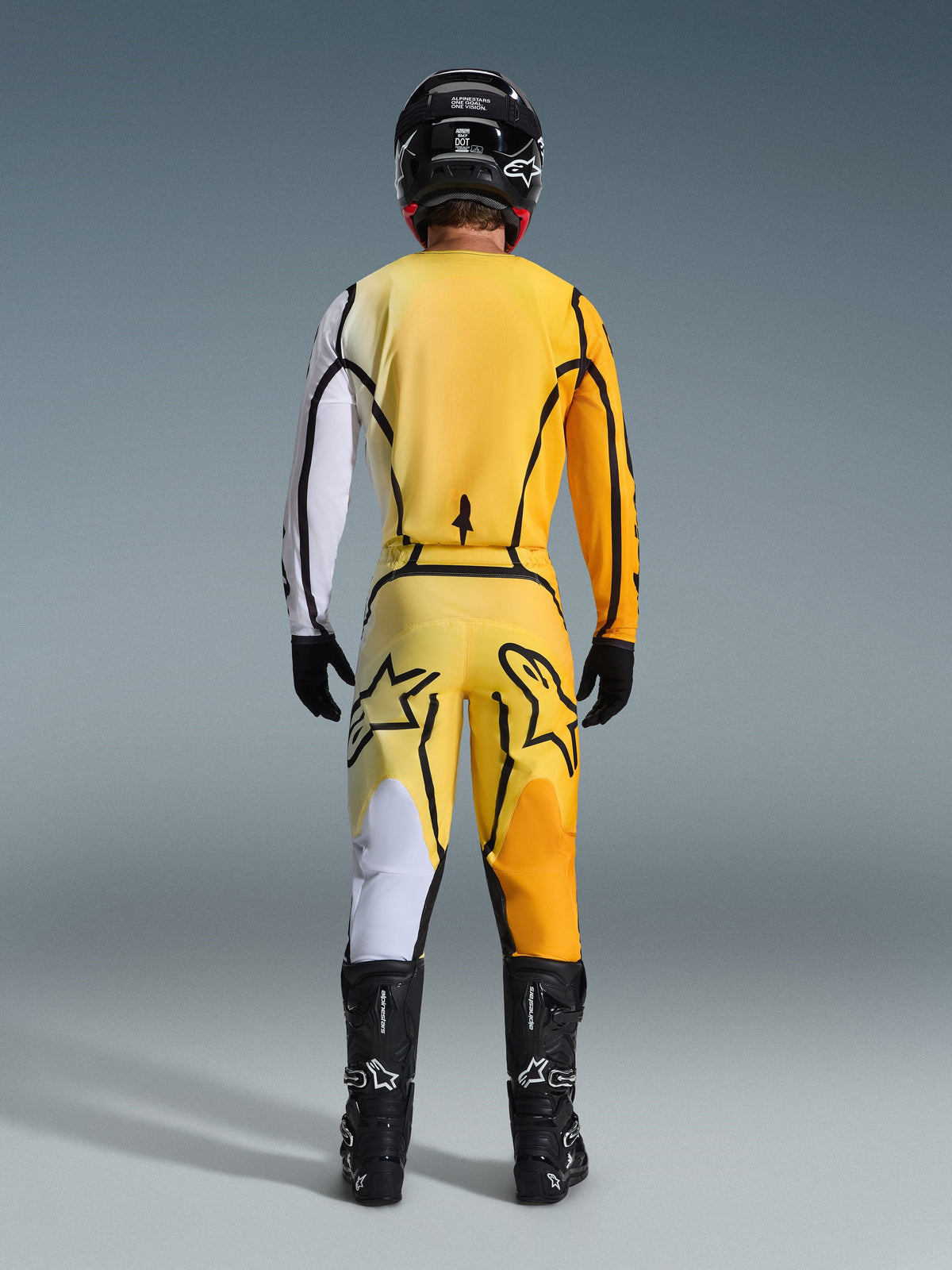 Alpinestars 2026 Fluid Apex crossbroek, motorcross broek, geel en zwart, achteraanzicht van een rijder in volledige motorcross-uitrusting, bijpassende jersey met witte en oranje kleurverlopen, zwarte helm, zwarte laarzen en zwarte handschoenen, gecoördineerde off-road outfit.