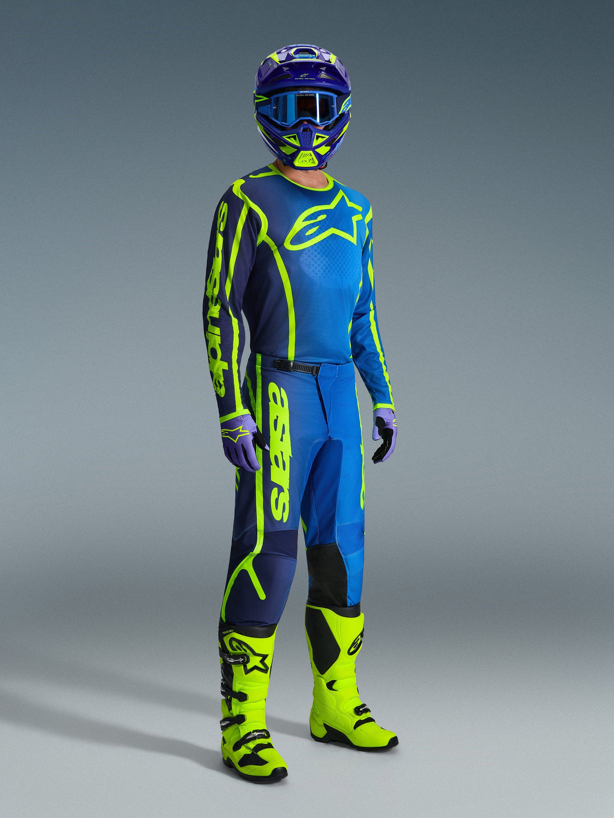 Pilote de motocross portant un pantalon Alpinestars Fluid Apex 2026 bleu et jaune fluo, coordonné avec un maillot assorti, un casque bleu et jaune et des bottes jaune vif, ensemble d'équipement professionnel de motocross.