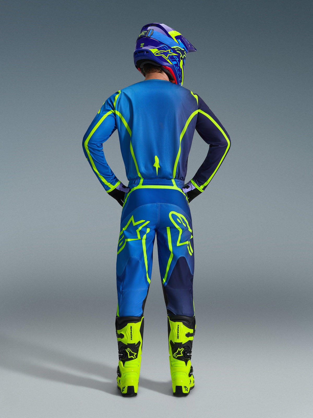 Alpinestars 2026 Fluid Apex Pants, pantalon de motocross, bleu et jaune fluo, vue arrière du pilote en tenue de course complète, maillot et casque assortis, bottes jaune fluo, coupe athlétique coordonnée pour le motocross.