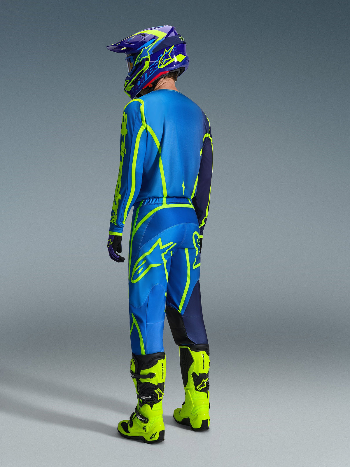 Pilote de motocross vu de dos portant le pantalon Alpinestars Fluid Apex 2026 bleu et jaune fluo, maillot et casque assortis, et bottes jaune fluo, équipement de course coordonné pour la performance tout-terrain.