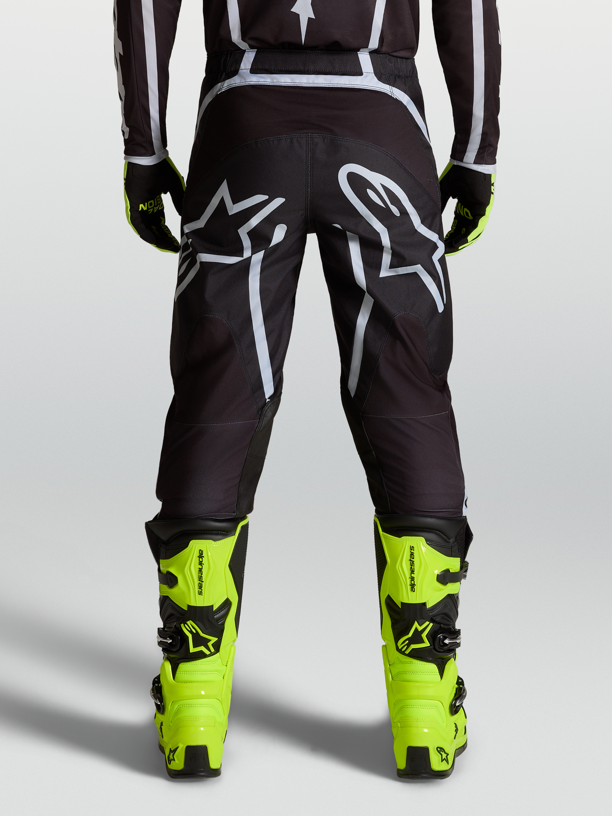 Alpinestars Pantalon Fluid Apex V2, pantalon de motocross noir et argenté, vue de dos sur un pilote avec bottes jaunes, coupe ergonomique avec panneaux extensibles et logo Astars sur le siège