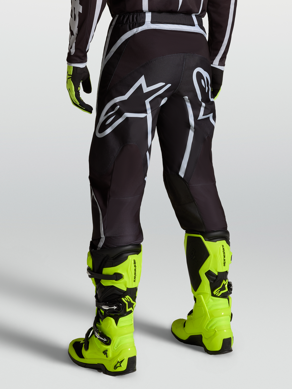 Alpinestars Pantalon Fluid Apex V2, pantalon de motocross noir et argenté pour homme, porté avec des bottes jaune fluo, vue arrière montrant le logo Astars et la coupe ergonomique en poly-tissu.