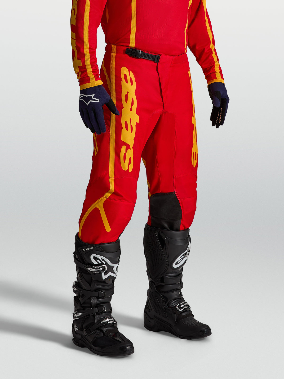 Alpinestars Fluid Apex V2, pantalon de motocross pour homme, rouge et doré, conçu en poly-tissu 600D robuste pour le tout-terrain, porté par un pilote avec des gants et bottes de moto noires.