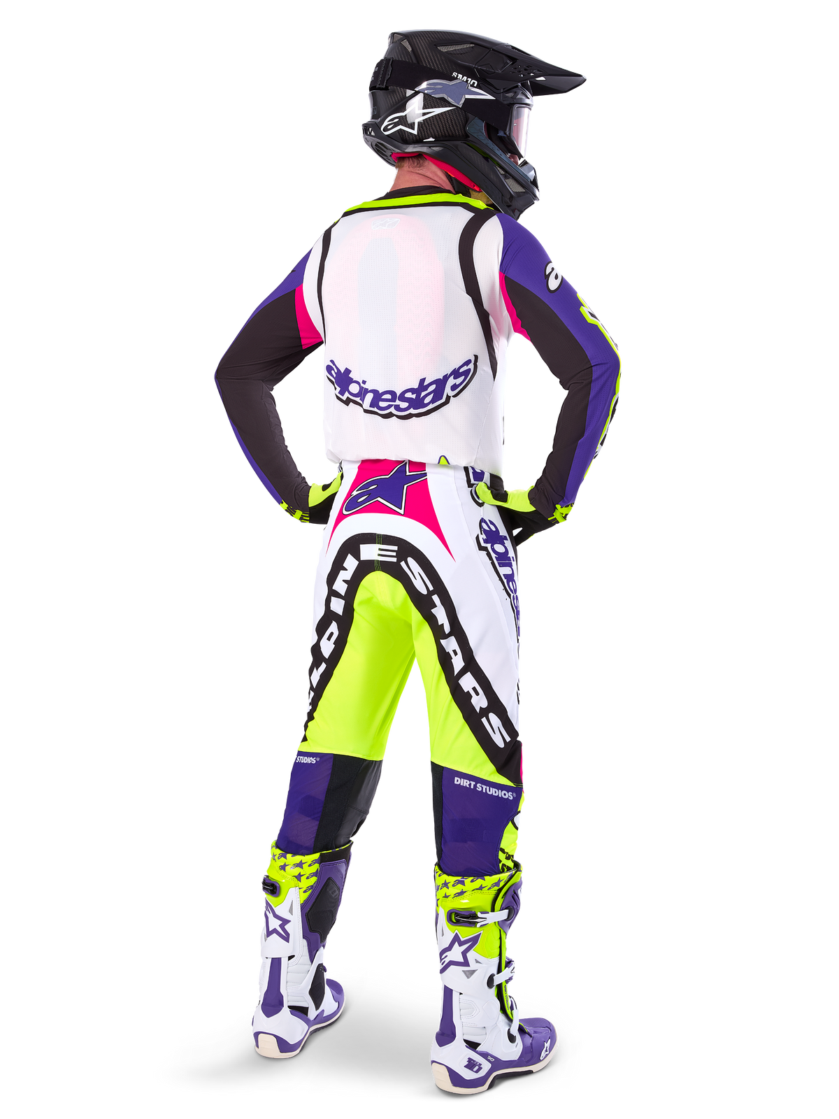Motocross-Fahrer in Alpinestars Limited Edition Supertech Dirt Studios® Hose in Weiß, Lila und Gelb Fluo, Rückansicht mit passendem Trikot, Stiefeln und schwarzem Carbon-Helm, professionelle Offroad-Rennausrüstung mit markanten Grafiken und Neon-Akzenten.
