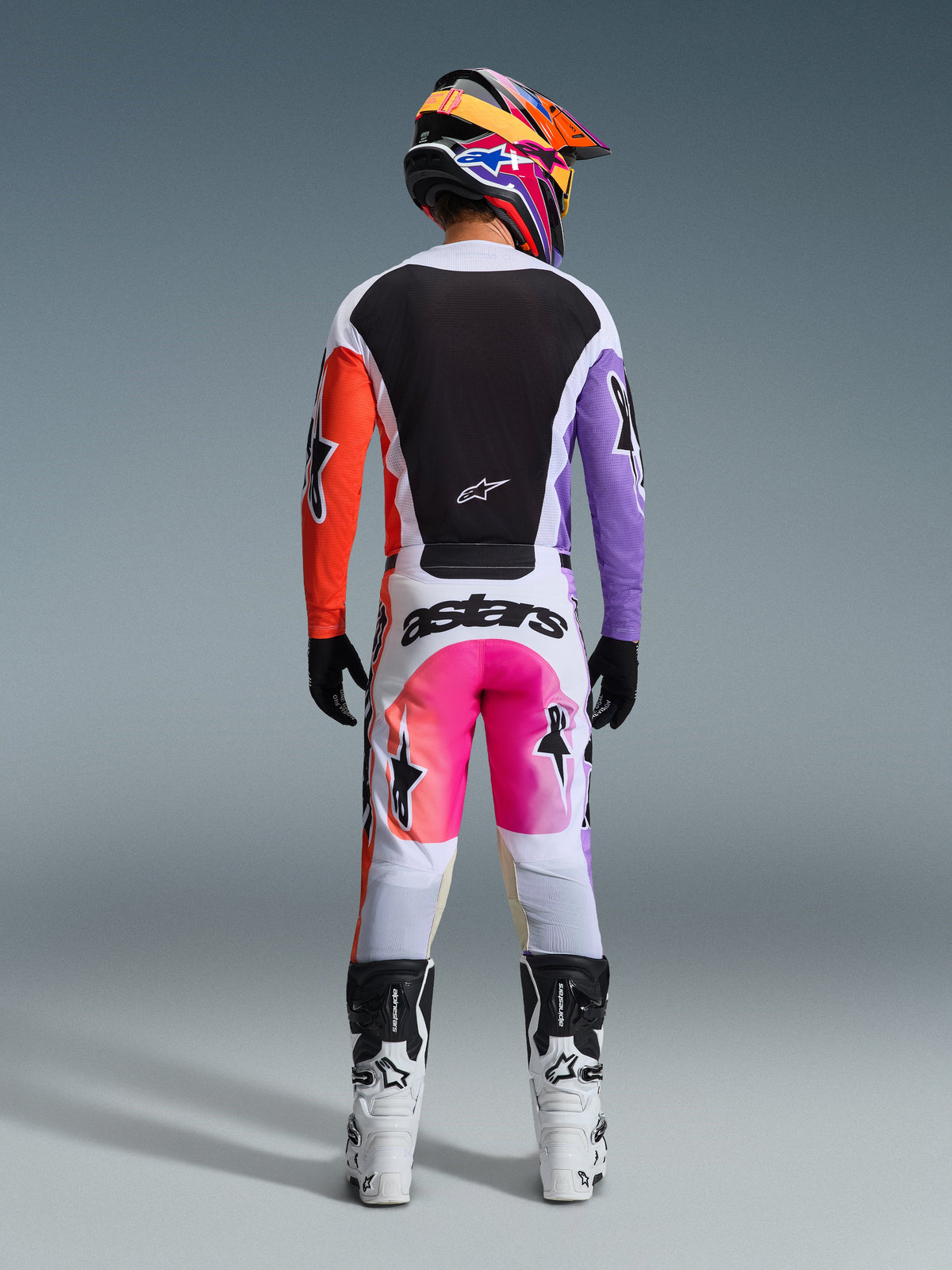 Pilote de motocross portant une tenue de course complète vue de dos, maillot Alpinestars 2026 Racer Air Portl avec empiècements blancs, noirs, orange et violets, pantalon blanc assorti avec touches de rose et logo 'astars', bottes blanches et noires, et un casque multicolore vif, tenue coordonnée pour le motocross