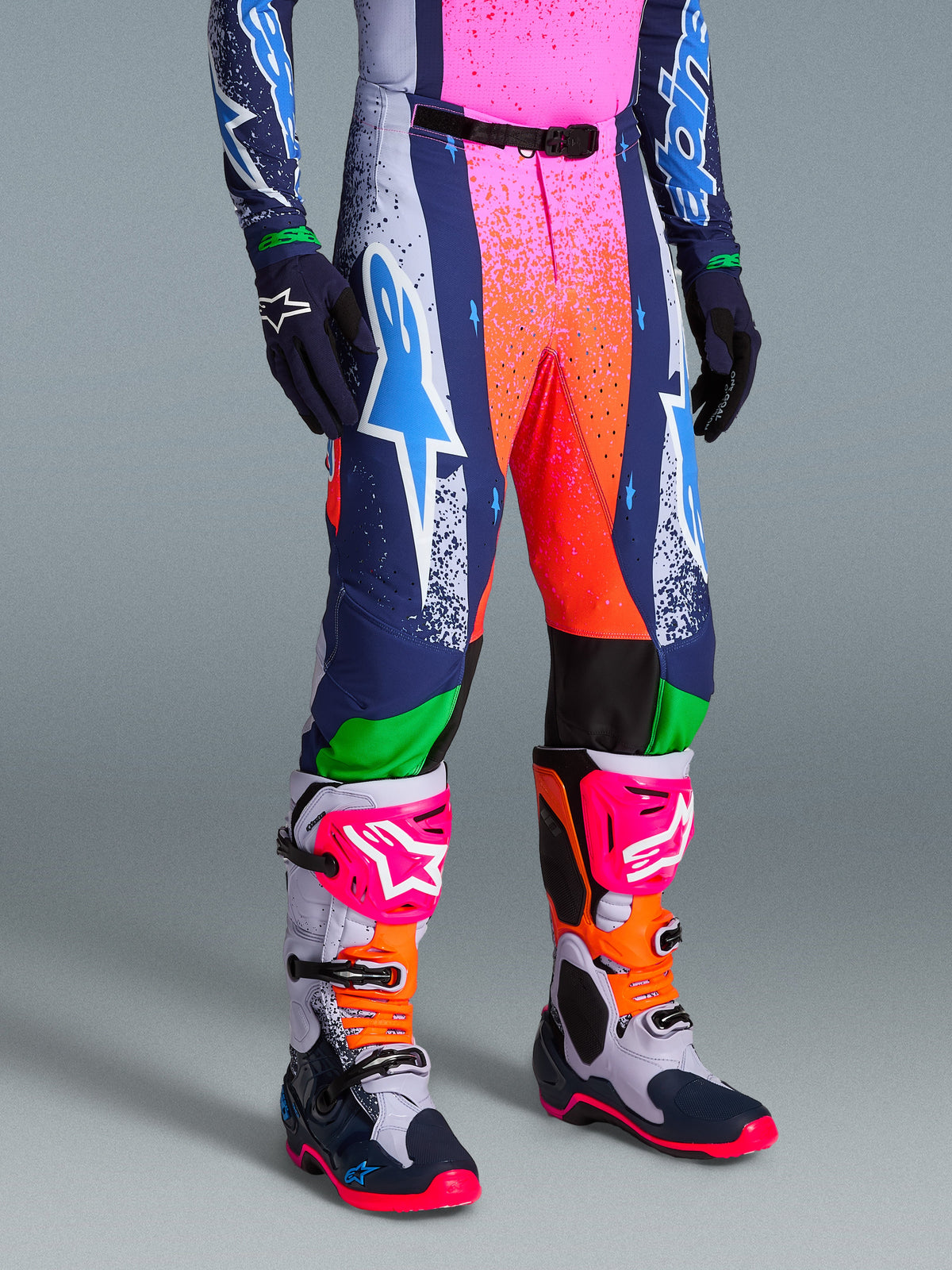 Alpinestars Limited Edition Supertech Records Hose, lebhafte mehrfarbige Motocross-Hose mit gesprenkelten Grafiken in Pink und Orange, dunkelblauen und weißen Einsätzen, mit einem großen weißen und cyanfarbenen Astars-Logo, getragen mit farblich abgestimmten Performance-Stiefeln und Handschuhen.