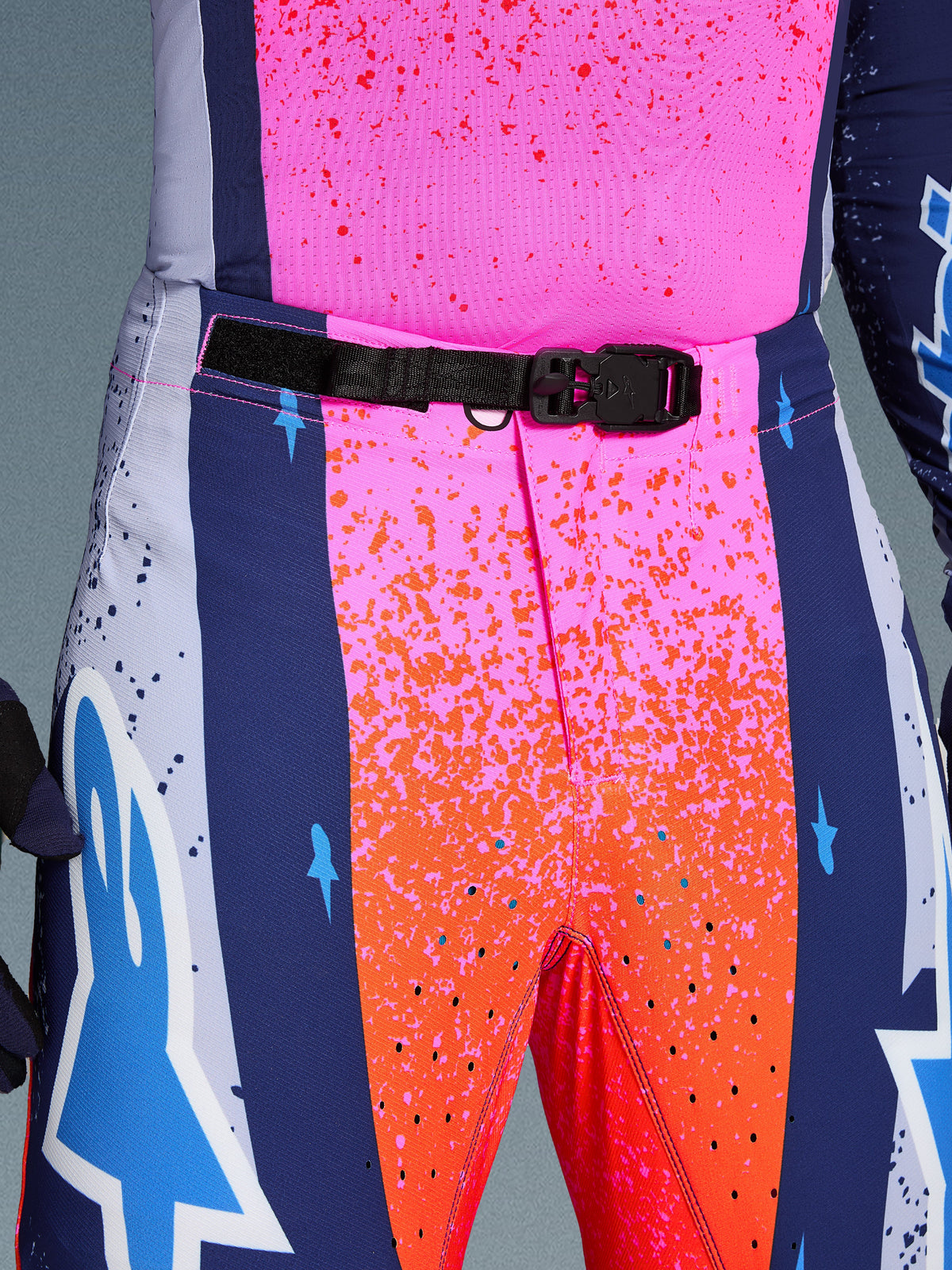 Alpinestars Limited Edition Supertech Records Hose, Motocross-Hose, Mehrfarbig Pink und Orange mit marineblauen Seiteneinsätzen, Nahaufnahme des Hosenbundes mit schwarzer, selbstverriegelnder SX1-Magnetschnalle, Ripstop-Gewebe mit Splatter-Grafiken und lasergeschnittenen Perforationen.