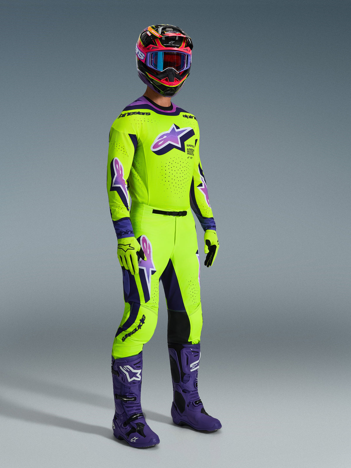 Ensemble d'équipement Alpinestars Supertech Vista 2026, pilote de motocross professionnel portant un maillot et un pantalon vert citron éclatant avec des accents et des logos violets, gants vert citron assortis, bottes violettes et casque multicolore avec lunettes bleues, tenue de course tout-terrain coordonnée