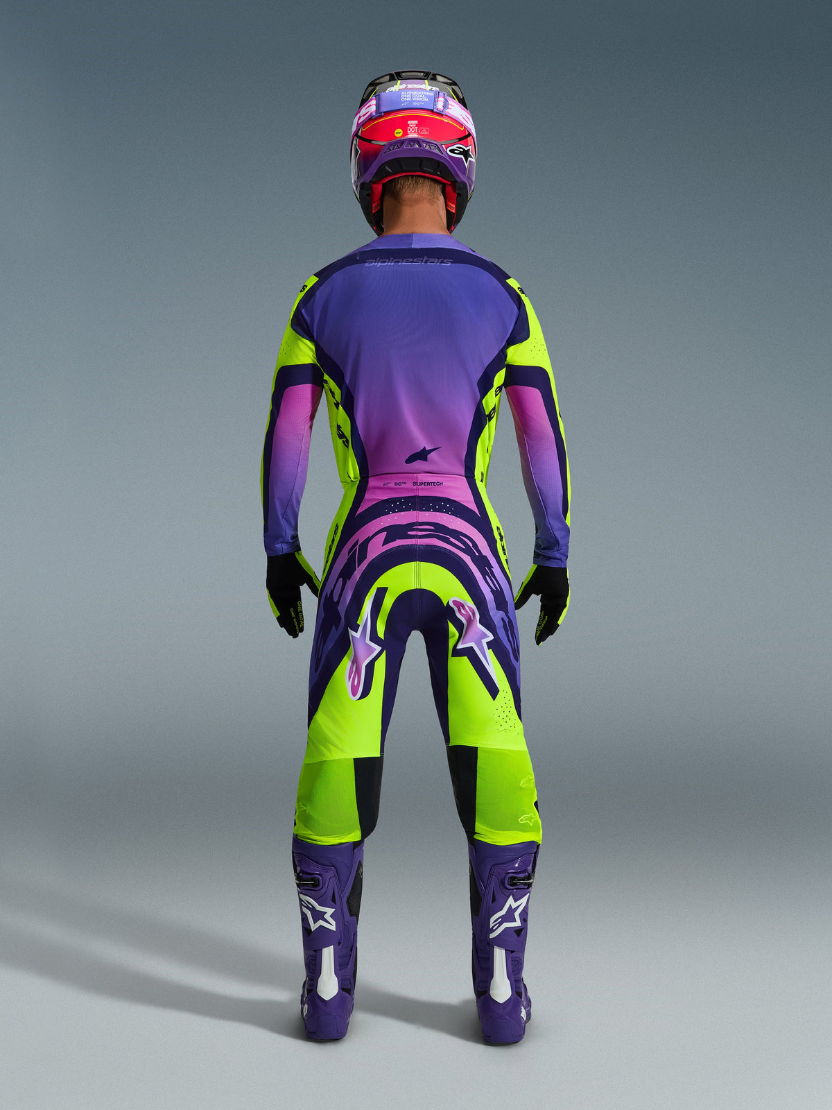 Ensemble d'équipement Alpinestars Supertech Vista 2026, vue arrière d'un pilote de motocross portant une tenue de course aux dégradés éclatants de violet, rose et vert citron, comprenant un maillot et un pantalon assortis avec un marquage audacieux, un casque coordonné violet et rose, des bottes violettes et des gants noirs.