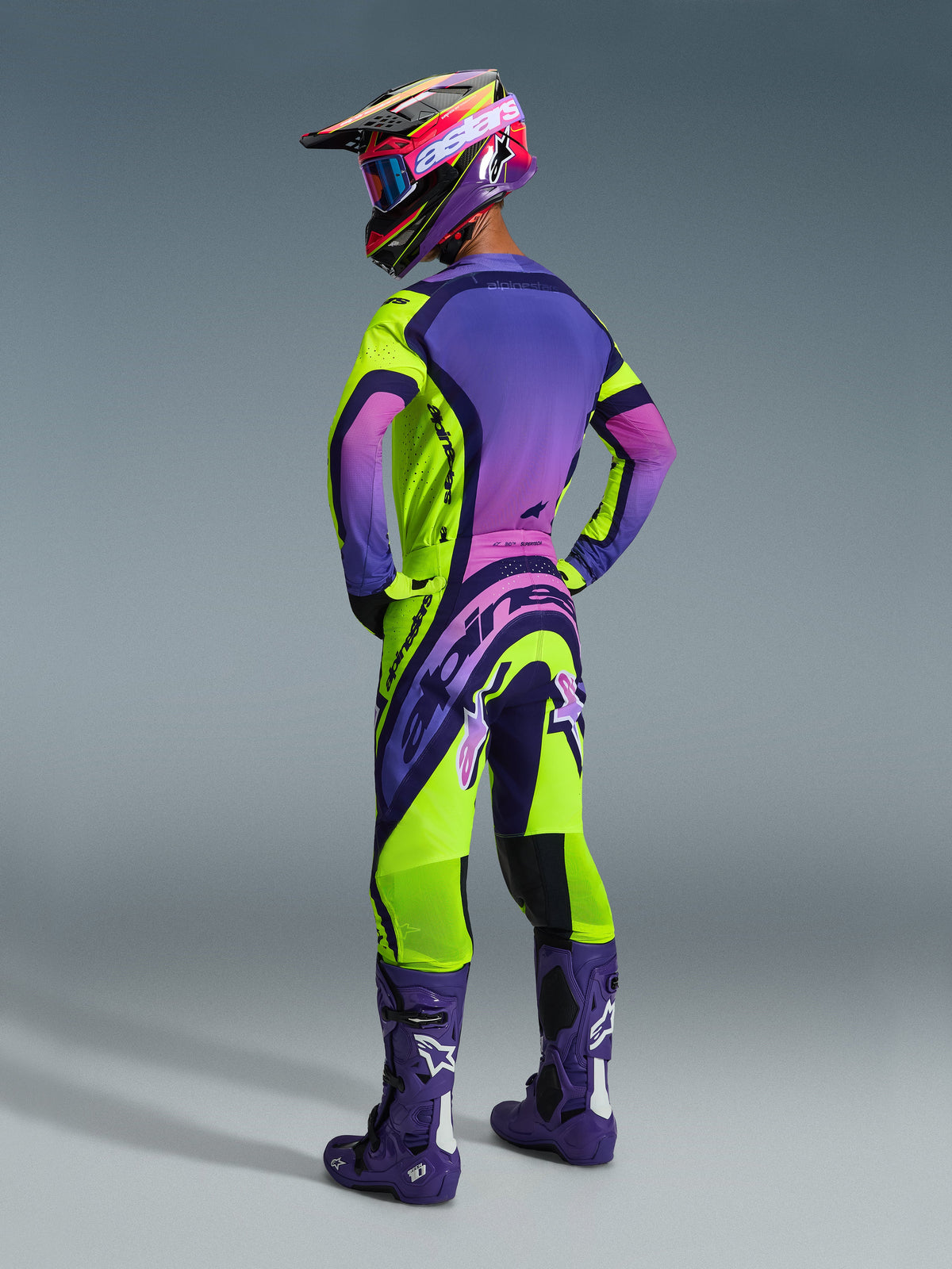 Pilote de motocross de dos portant l'ensemble Alpinestars Supertech Vista 2026, maillot violet avec dégradé rose et accents jaune fluo, pantalon assorti jaune fluo et violet, bottes violet foncé avec logos blancs, casque et lunettes coordonnés, équipement de course de motocross professionnel