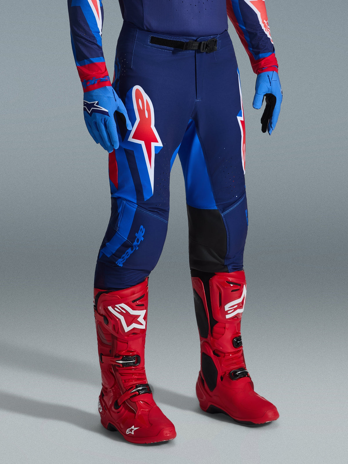 Alpinestars 2026 Supertech Vista Pants, Motocross Pants, Dark Navy Hot Coral Cobalt, worn with bright red racing boots and blue gloves, featuring large graphic logos on the thighs and black reinforced inner knee panels for off-road performance.