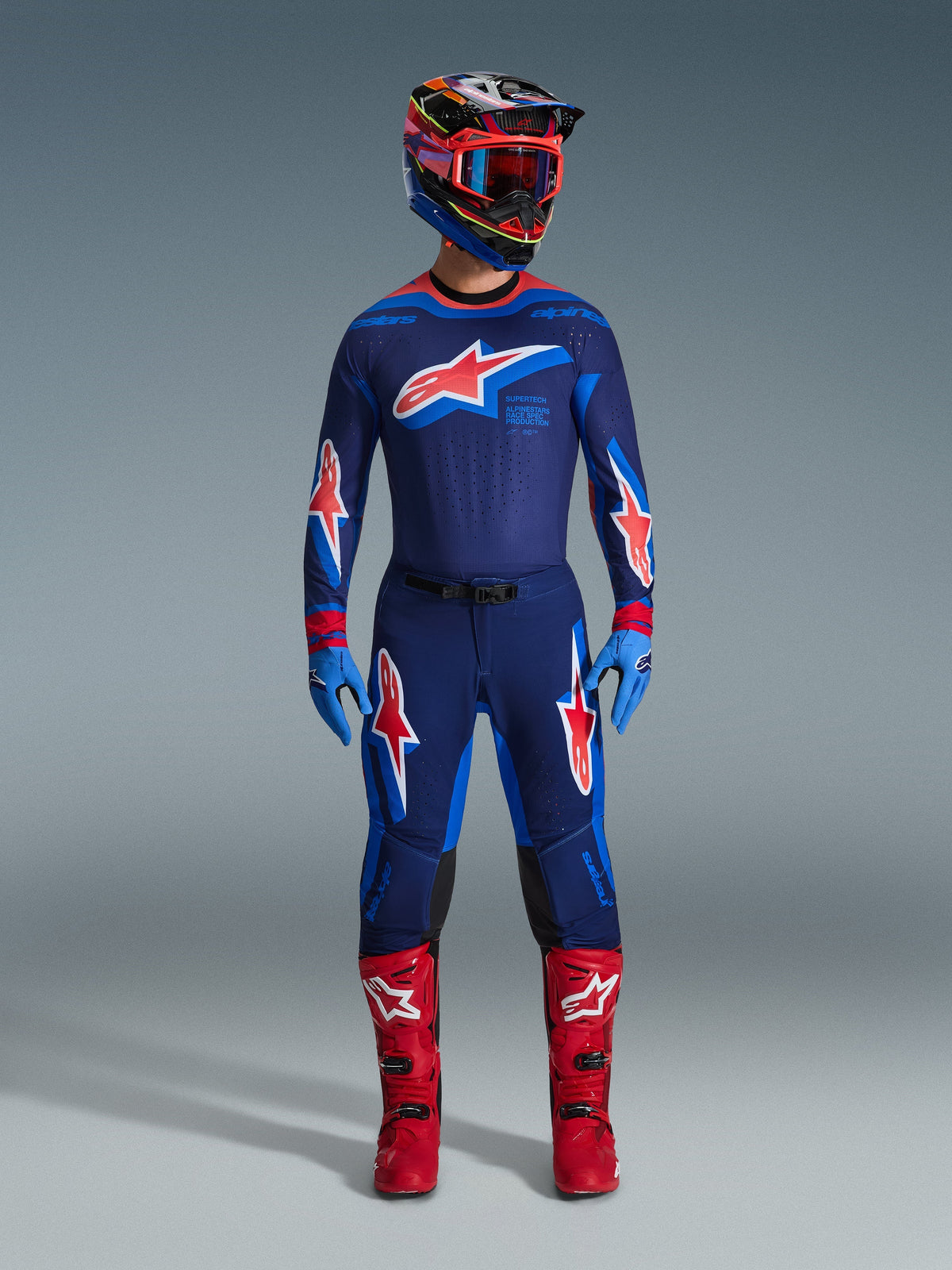 Motocross rider wearing complete racing gear, Alpinestars 2026 Supertech Vista Pants in Dark Navy Hot Coral Cobalt, matching jersey with prominent star logo, bright red boots, and helmet with red goggles, coordinated professional kit for off-road performance