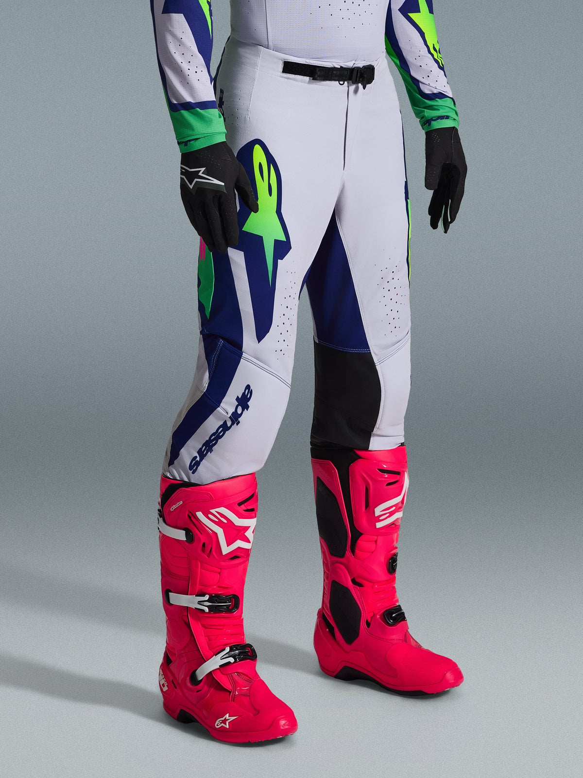 Alpinestars 2026 Supertech Vista broek, motorcrossbroek, lichtgrijs met fluogeel en blauwe accenten, gedragen met felroze motorcrosslaarzen en zwarte handschoenen, atletische racepasvorm met duurzame stretchconstructie voor elite off-road prestaties