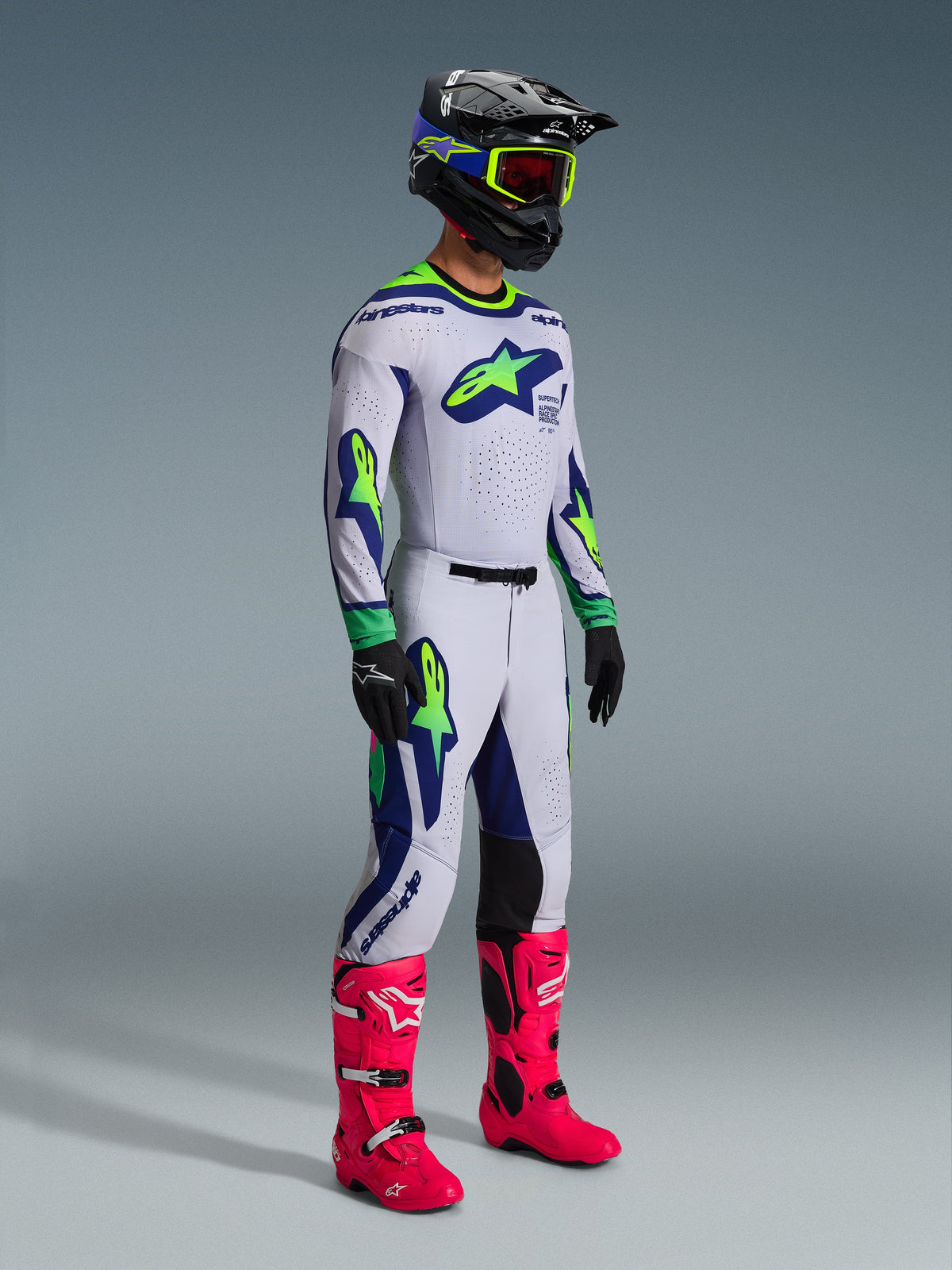 Ensemble d'équipement Alpinestars Supertech Vista 2026, pilote de motocross en tenue de course complète, maillot et pantalon blancs avec accents vert citron et bleu marine, bottes rose vif, casque noir avec détails bleus et vert citron, équipement tout-terrain coordonné de niveau professionnel