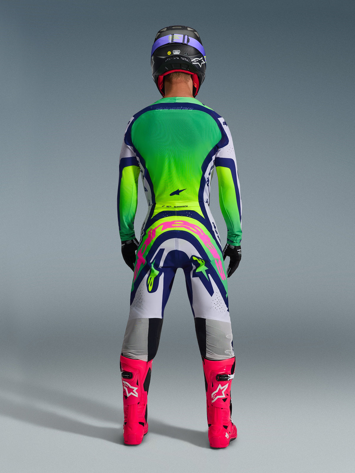 Ensemble d'équipement Alpinestars Supertech Vista 2026, vue arrière d'une tenue de motocross, maillot avec dégradé vert et jaune, pantalon blanc et bleu avec un grand logo rose à l'arrière, bottes rose vif et casque violet, tenue de course tout-terrain professionnelle
