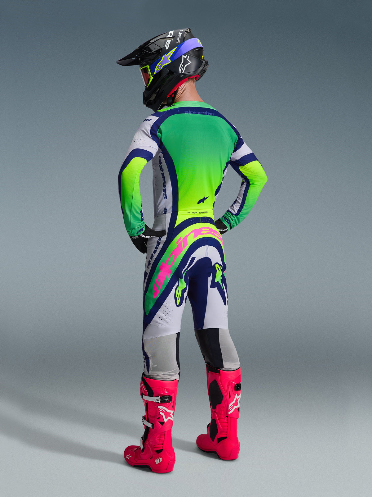 Vue arrière d'un pilote de motocross portant l'ensemble d'équipement Alpinestars 2026 Supertech Vista, maillot blanc et vert citron avec des accents bleus, pantalon assorti avec un logo rose vif à l'arrière, bottes de course rose vif et casque noir avec lunettes vert citron, tenue de course tout-terrain professionnelle