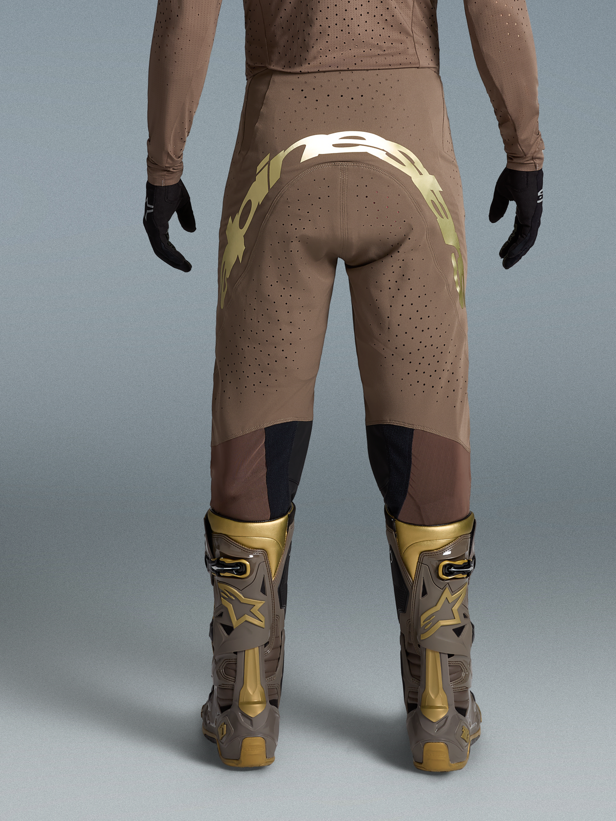 Alpinestars Beperkte Oplage Supertech Air Gold broek, bruine en gouden motocrossbroek met geperforeerd textiel en bijpassende Tech 10 laarzen op een model, achterzicht.