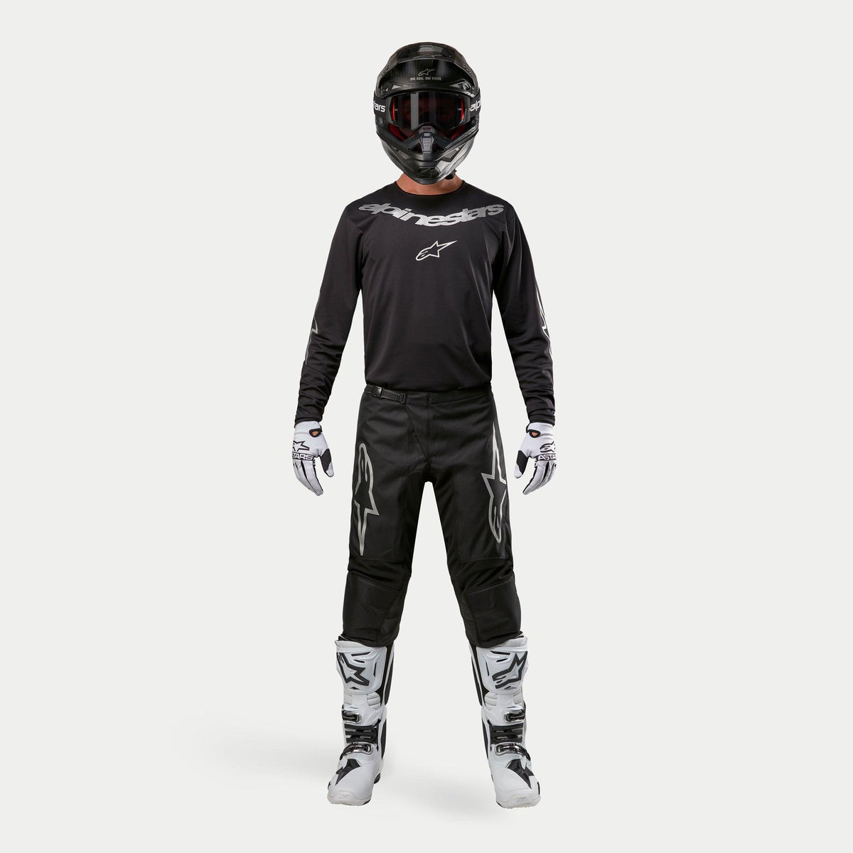 Motocross-Fahrer in kompletter Rennausrüstung, Alpinestars 2024 Fluid Graphite Hose in Schwarz und Silber, schwarzes Langarm-Trikot mit silbernem Branding, schwarzer Helm mit Goggles, weiß-schwarze Stiefel und Handschuhe, abgestimmtes Outfit für Motocross