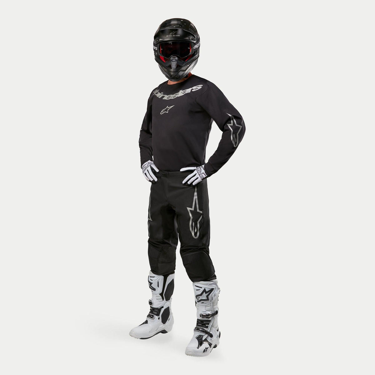 Alpinestars 2024 Fluid Graphite Hose, Motocross-Hose, Schwarz und Silber, getragen mit passendem schwarzem Jersey, schwarzem Helm, weißen Handschuhen und weißen Stiefeln, silberne Alpinestars-Logos auf den Oberschenkeln, ergonomische athletische Passform für den Offroad-Rennsport