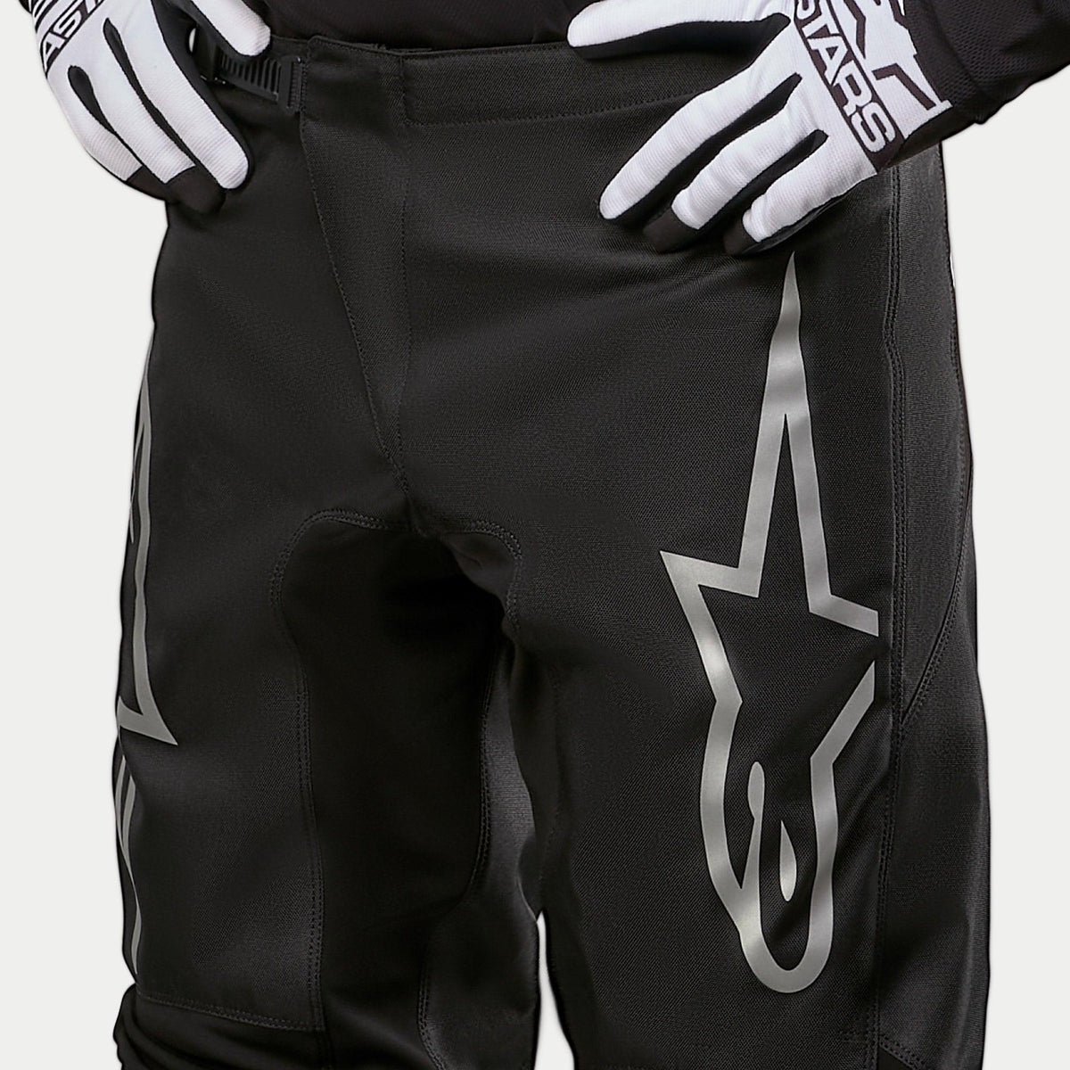 Alpinestars 2024 Fluid Graphite Hose, Motocross-Hose, Schwarz und Silber, Detailaufnahme der Oberschenkel und Taille, mit einem großen silbernen Stern-Logo auf dem Bein, getragen mit schwarz-weißen Rennhandschuhen