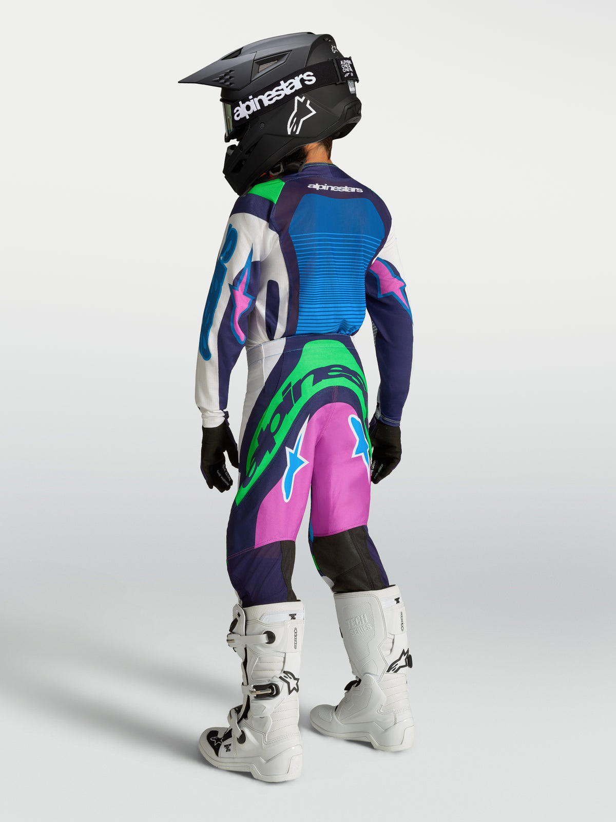 Alpinestars Jeugd Supertech Maven broek, motocrossbroek voor jonge rijders, blauw, roze en groen, atletische racefit, afgebeeld met witte motorlaarzen.