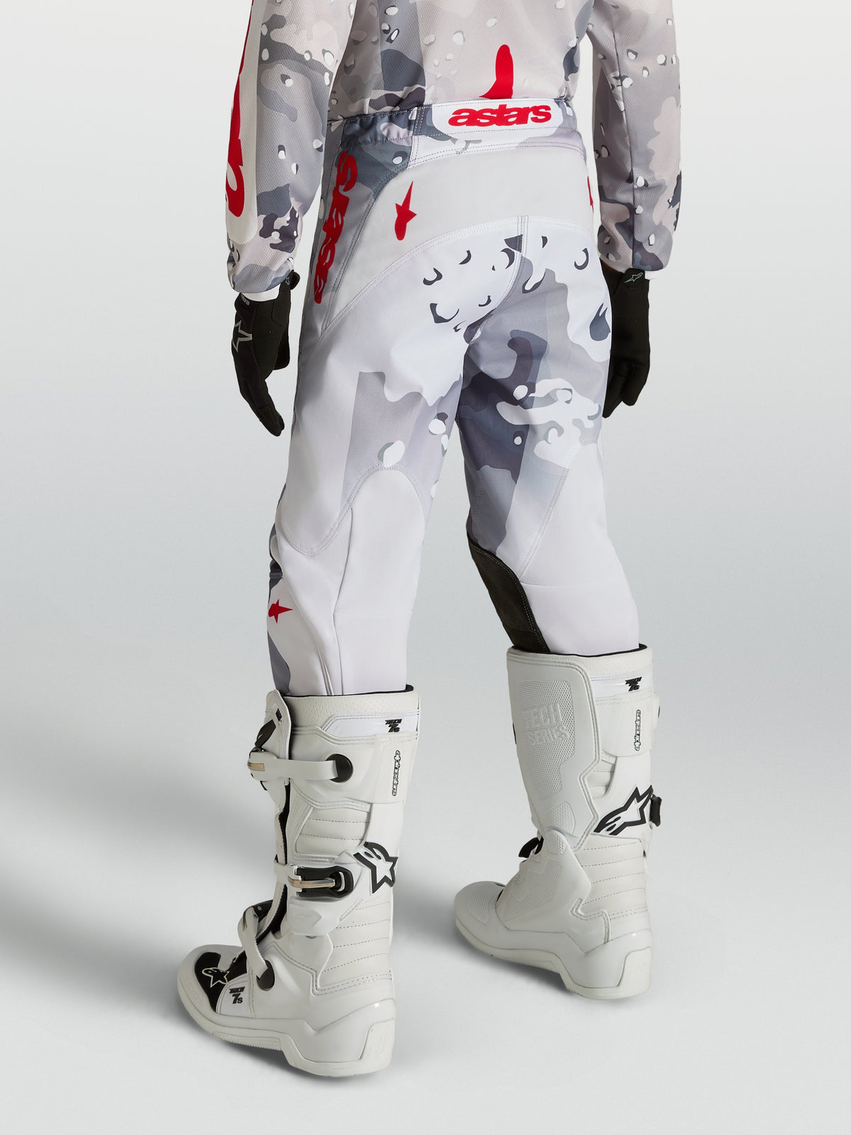 Pantalon Fluid Lone Junior d'Alpinestars, pantalon de motocross tout-terrain pour enfant, camouflage gris et rouge, coupe ergonomique en poly-tissu durable, porté avec des bottes blanches.