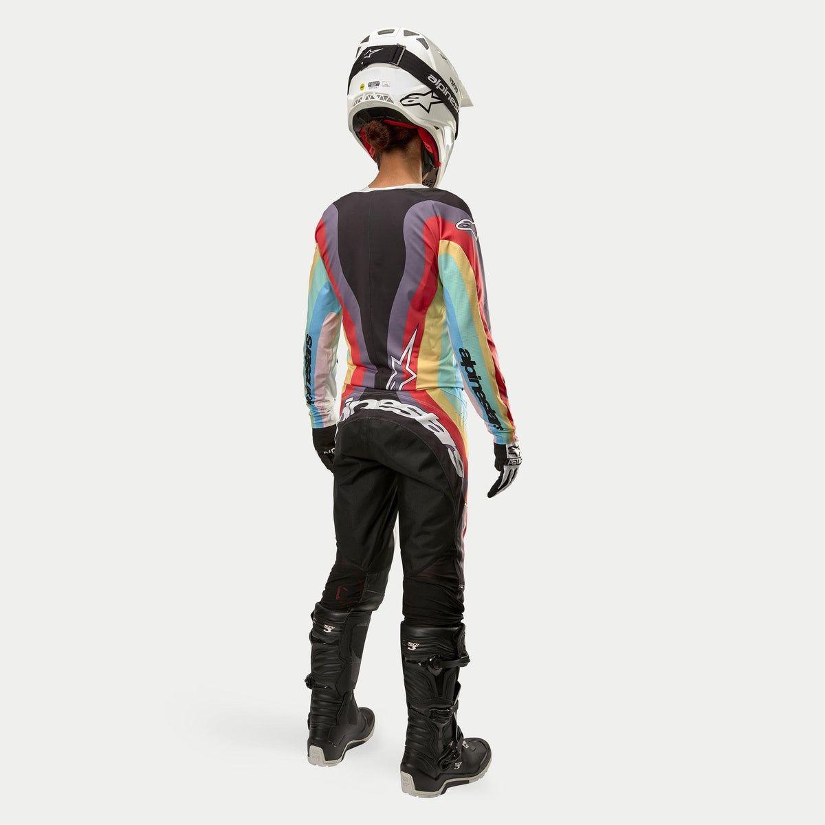 Motocross rider from behind wearing Alpinestars Women Stella 2024 Techstar Pants in Black Multicolor, featuring large white branding on the seat, coordinated with a colorful striped jersey, white helmet, and black Tech 3 boots for off-road racing.