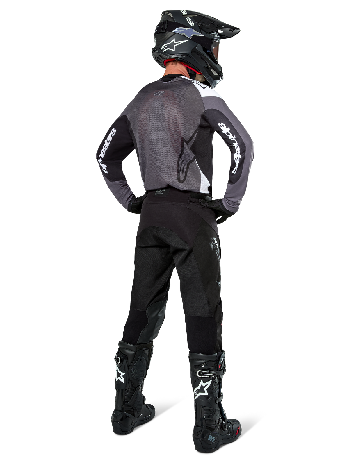 Pilote de motocross portant le maillot Alpinestars Pro-Dura en Black Magnet White, vue arrière montrant le panneau arrière en maille grise et les manches noires avec logo Alpinestars blanc, coordonné avec un pantalon noir, un casque noir et des bottes noires et blanches, équipement de course d'enduro professionnel
