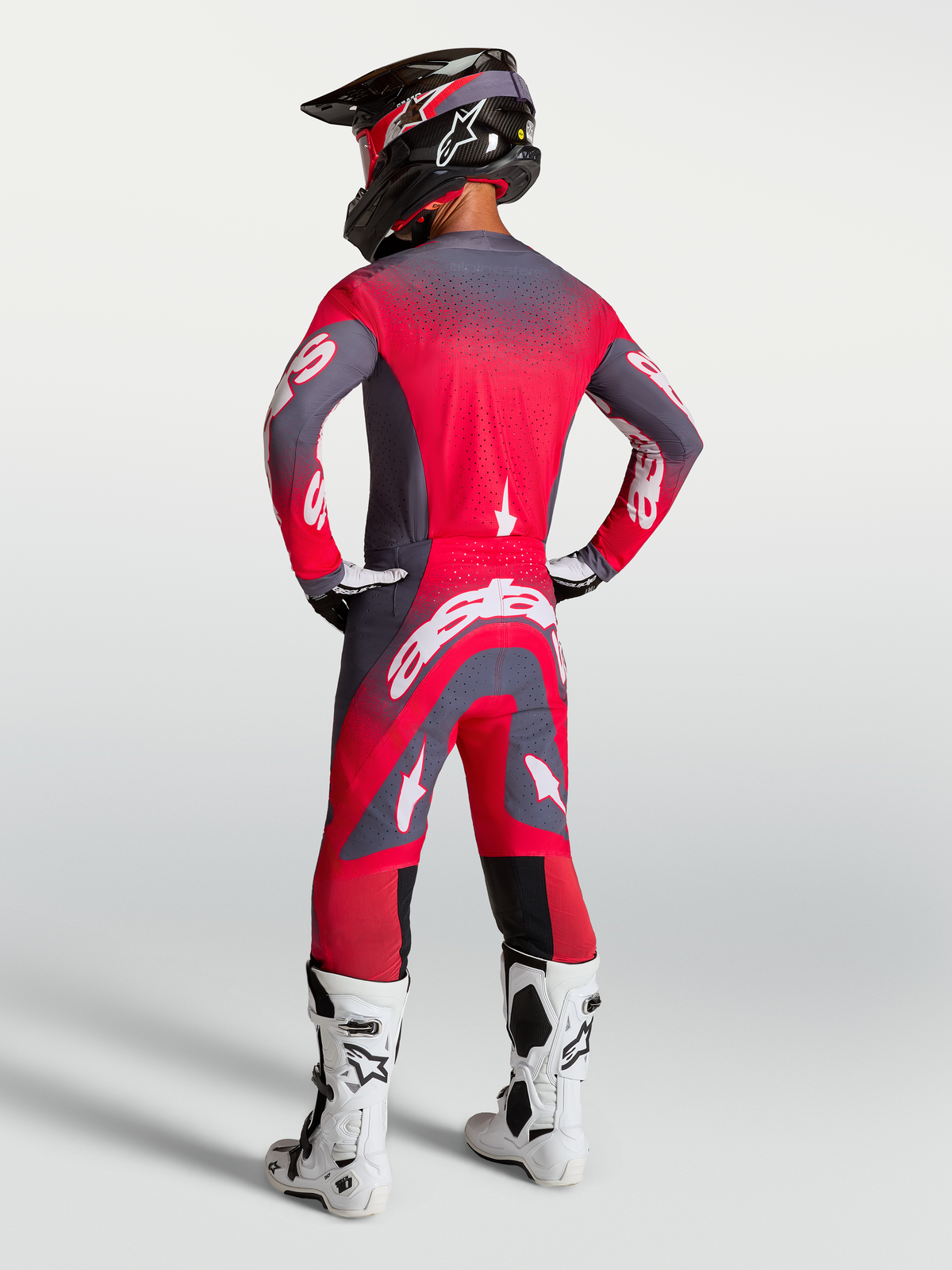 Alpinestars Supertech Hiten AFD Jersey en Gear Set, professionele motocross-uitrusting, rood en grijs, achteraanzicht van model met helm en witte motorlaarzen.