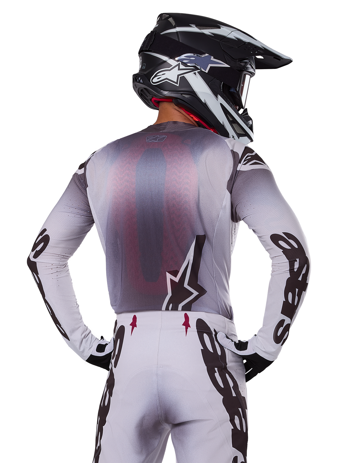 Alpinestars 2025 Supertech Maker Gear Set, motorcrosser van achteren gezien, wit en grijs shirt met grote zwarte merknaam op de mouwen en ster-logo, bijpassende witte broek met zwarte graphics, zwart-witte helm, professionele off-road race-outfit