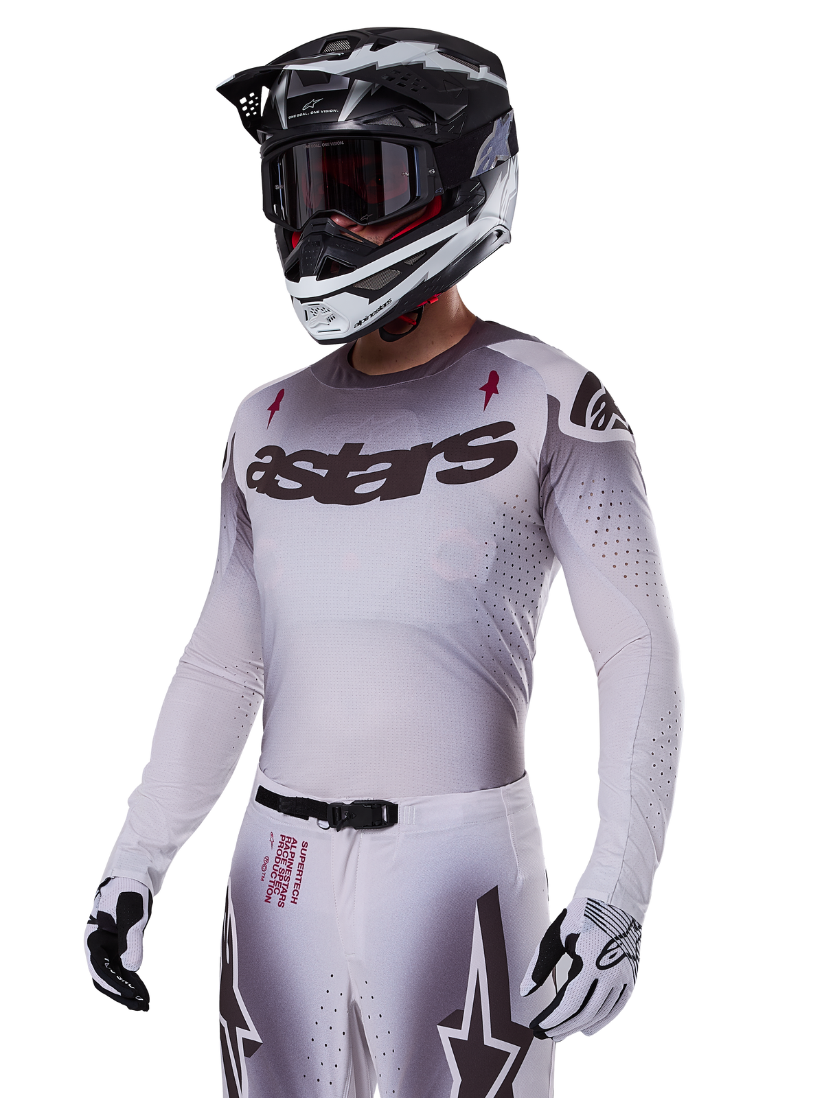 Motorcrosser die de Alpinestars 2025 Supertech Maker Gear Set in het wit draagt, met een jersey met grijs kleurverloop, het zwarte astars-logo en lasergesneden perforaties, bijbehorende broek met magnetische gesp en ster-graphics, gedragen met een zwart-witte helm en handschoenen voor een complete off-road race-look.